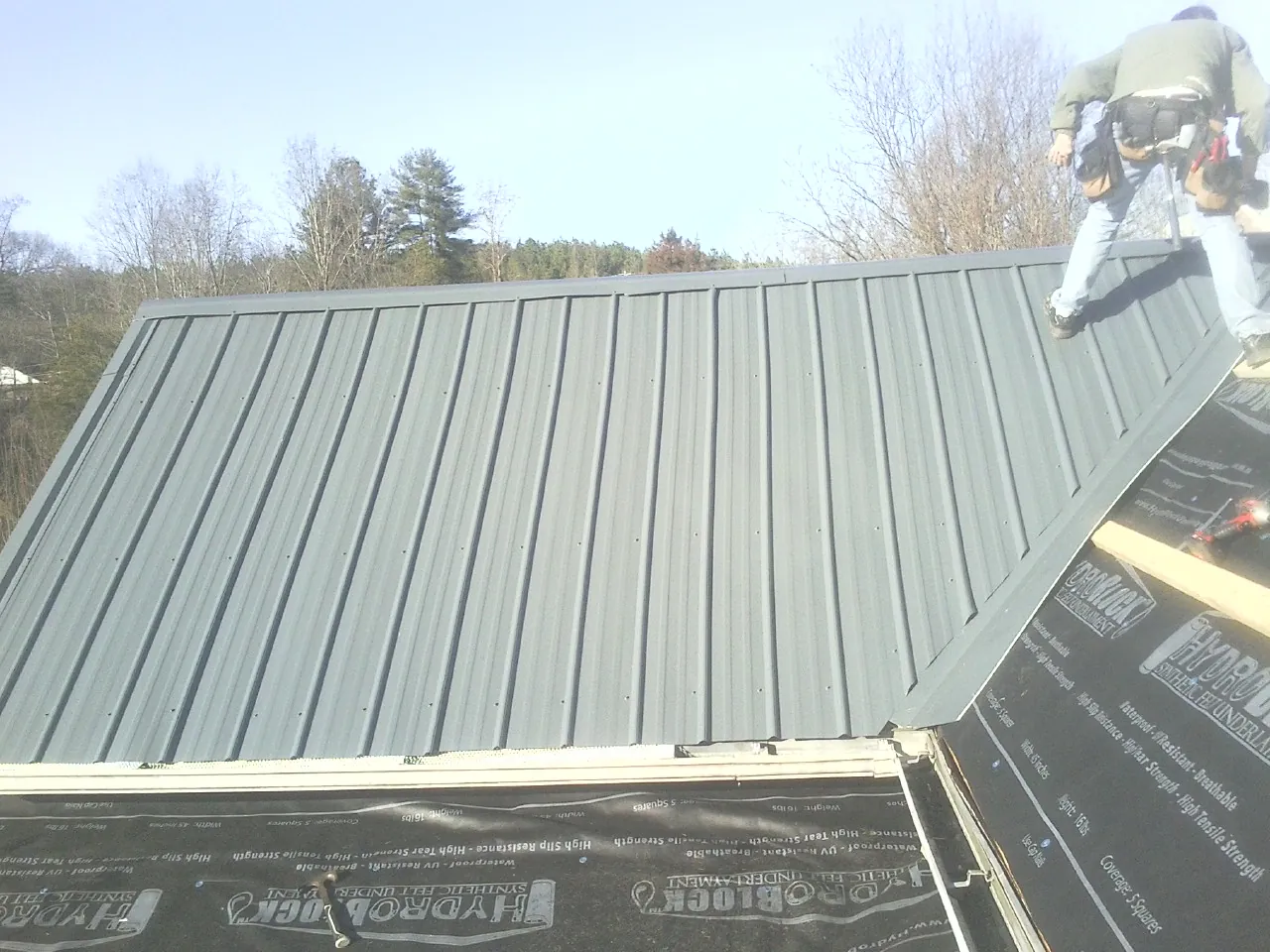 Crew installing dark green standing seam panels over synthetic underlayment in Portis