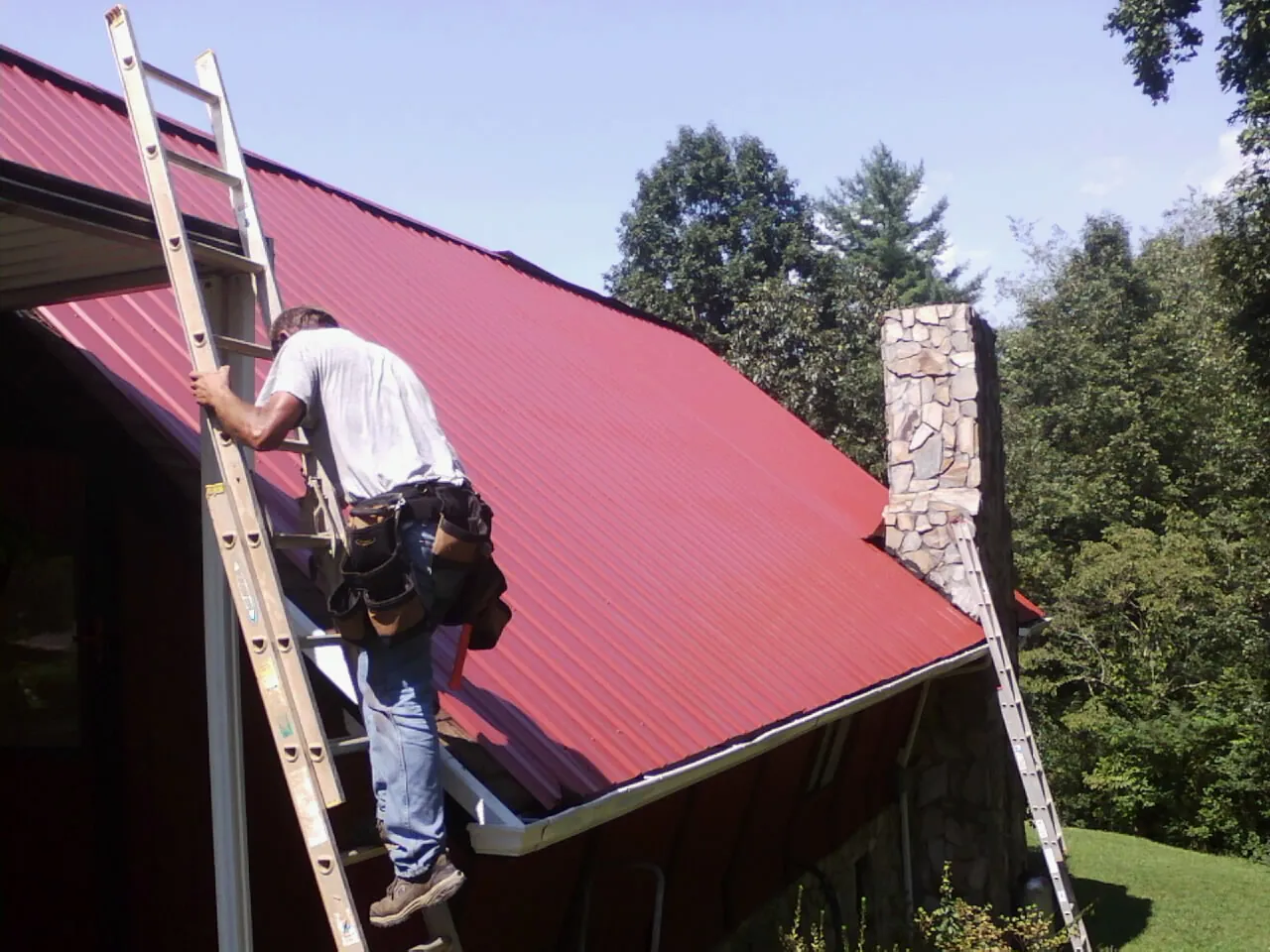 Roofer on a ladder beside a red metal roof and stone chimney — Soffit & Fascia Repair by Roofing Brothers in Portis